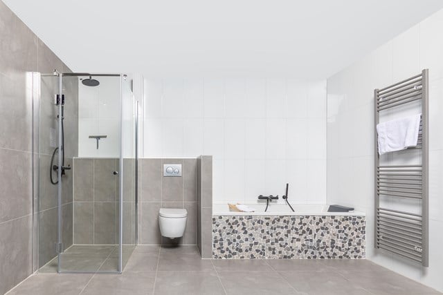 Moderne badkamer met glazen regendouche, wit toilet, en ligbad met mozaïekrand; stijlvol in grijze en witte tinten.