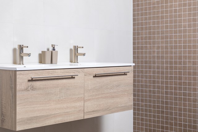 eiken-badkamermeubel-twee-lades.jpg Houten badkamermeubel met dubbele wastafel en moderne kraansets, afgewerkt in licht eiken, tegen een beige mozaïek wand.