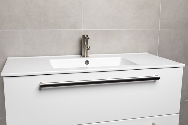 wit-badkamermeubel.jpg Witte badkamermeubel met geïntegreerde keramische wastafel, zwarte handgreep en een moderne kraan tegen een grijze tegelwand.