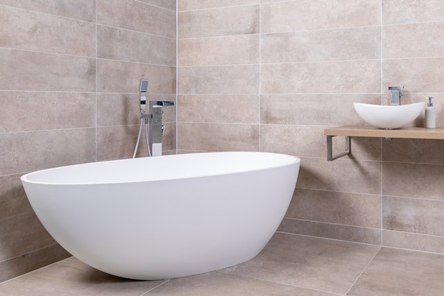 Ovale vrijstaande witte badkuip in modern beige badkamer, gemaakt van kunststof, met minimalistische design en metaalkleurige kraan.