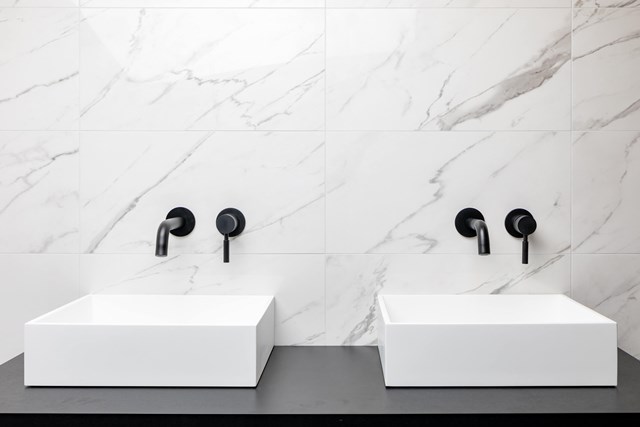 Zwei quadratische weiße Waschbecken auf einem Dunkelgrau Badmöbel, mit mattschwarzen Wandarmaturen gegen eine marmorierte Rückwand.
