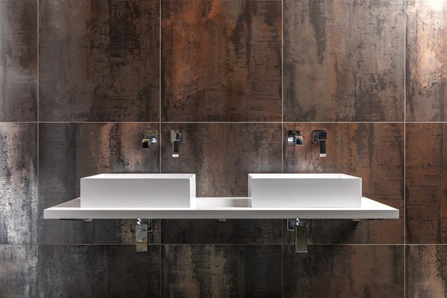 Weiße Doppelwaschbecken mit klarem Design auf einer schwebenden Platte, befestigt an einer Wand mit dunklen, rostfarbenen Fliesen.