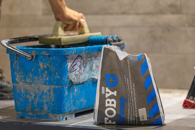 Blauw bouwemmer met handvat en zak grijze voegenmortel, ideaal voor badkamertegels en renovaties.