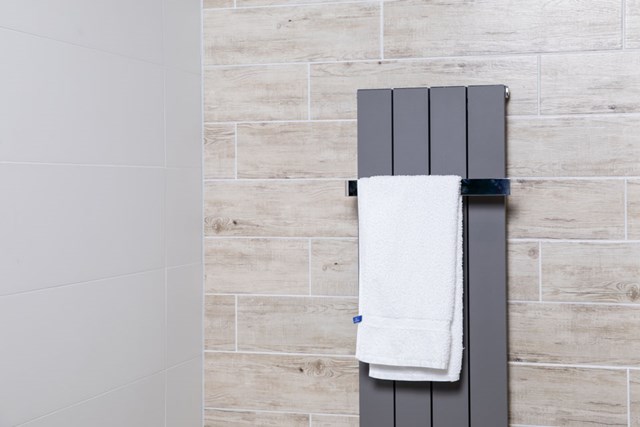 Grauer vertikaler Design-Heizkörper mit weißem Handtuch, wandmontiert in einem modernen Badezimmer mit warmer Holzoptik-Fliesenwand.