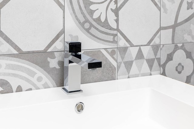 Viereckiger chromfarbener Badezimmerhahn mit schwarzen Details, platziert auf einem weißen Waschbecken mit grauen Fliesen als Hintergrund.