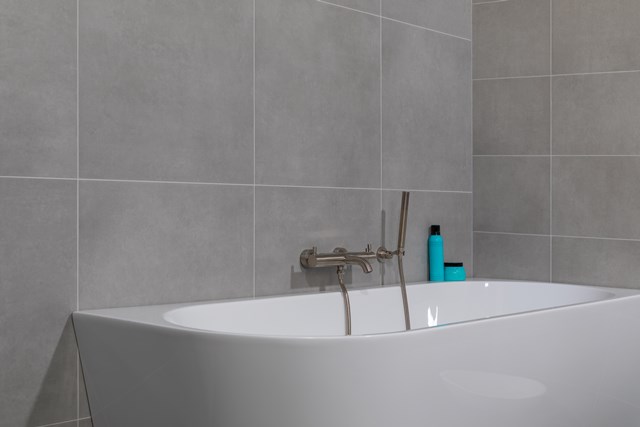 Weißes ovales Bad mit modernem Edelstahlarmatur und Handbrause, platziert gegen hellgraue Fliesen in einem stilvollen Badezimmer.