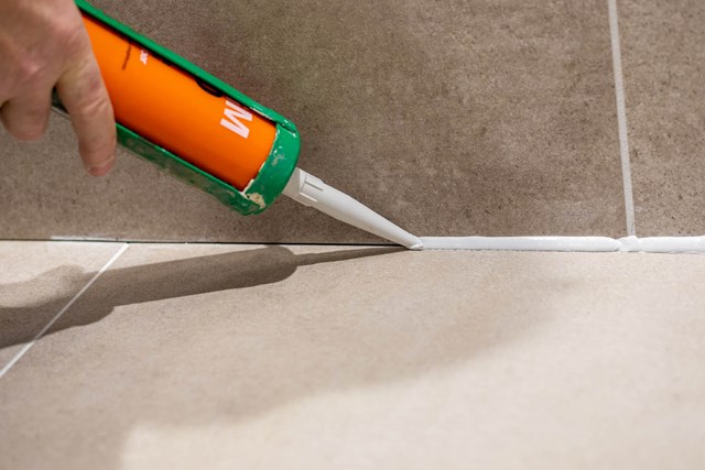 Oranje-en-groene kitpistool met witte siliconenkit, aangebracht op lichtgrijze tegels in een moderne badkamer.