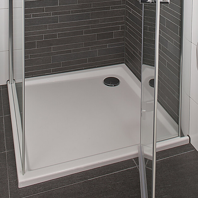 White square shower tray made of plastic with low entry, placed in a modern bathroom with dark tiles.