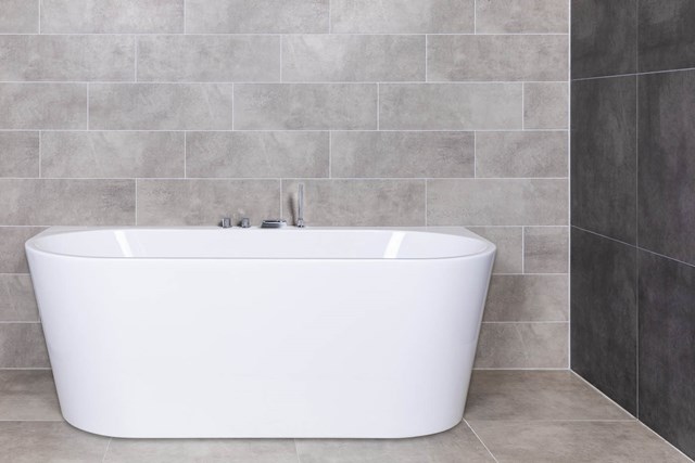 Vrijstaande witte badkuip van acryl met een modern design, geplaatst tegen een muur met grijze tegelafwerking in een badkamer.