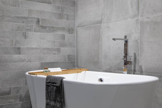 ideale-hoogte-bad-betonlook-tegels-vrijstaand-bad-vrijstaande-badkraan.jpg Wit vrijstaand bad met houten badplank en handdoek, geplaatst in een moderne badkamer met grijze tegelmuur.